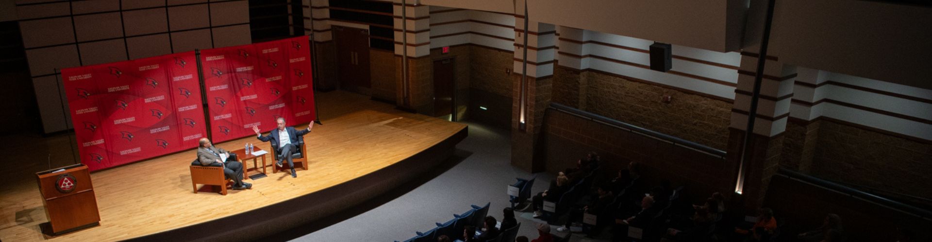 two people in chairs on a stage facing a crowd of viewers