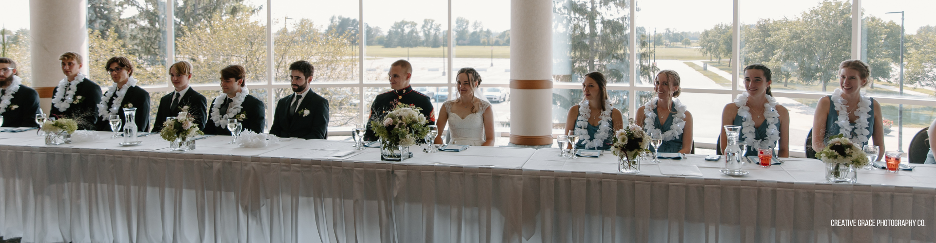 wedding party seated at a long rectangular table with floral centerpieces in front of a window