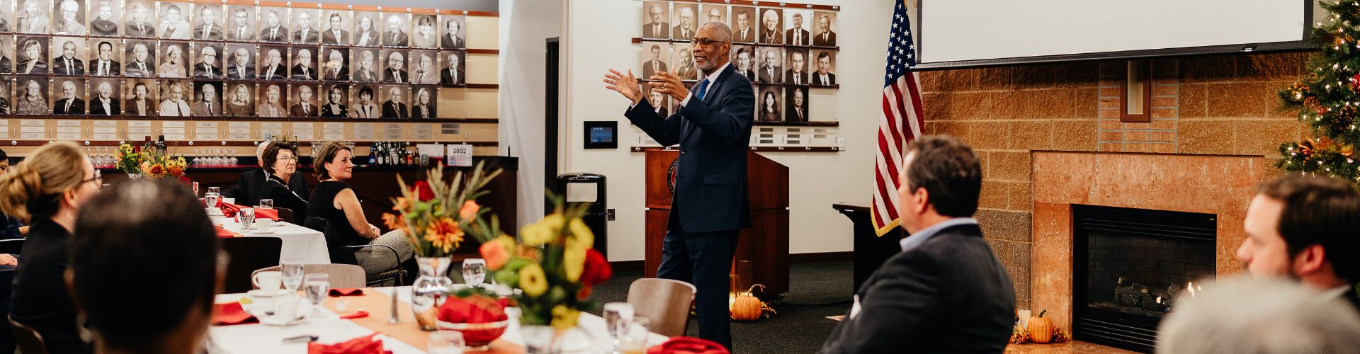 a man standing and speaking to seated listeners in a room decorated with floral centerpieces