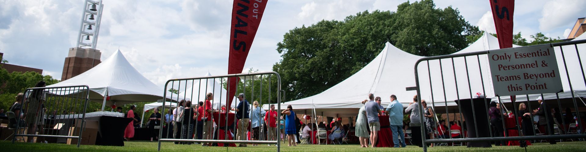 multiple white tents on a lawn in front of a bell tower with people standing nearby
