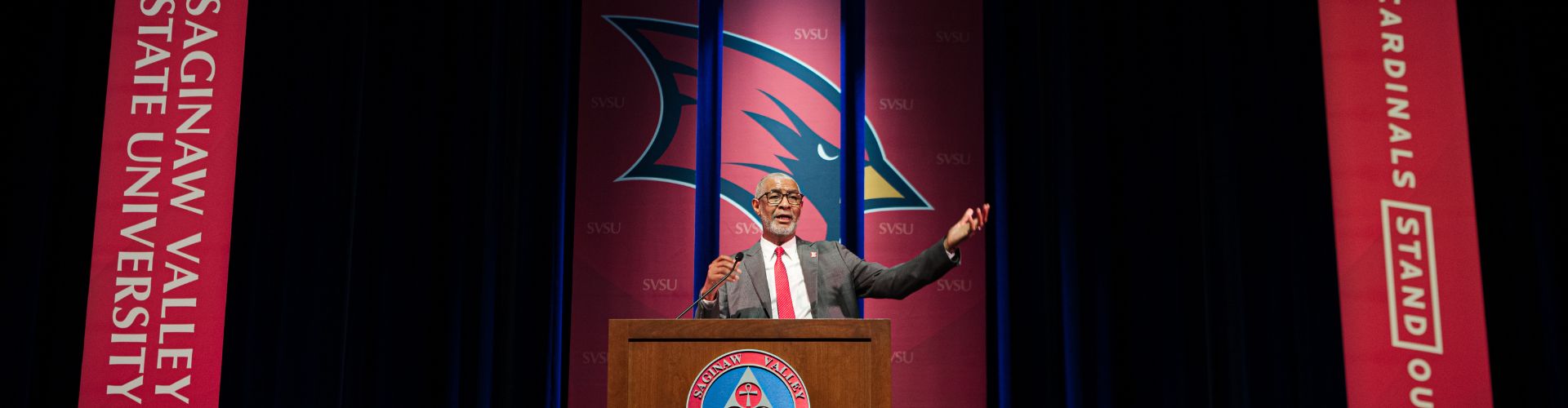 A man in a suit standing at a lectern speaking in front of large SVSU branded banners