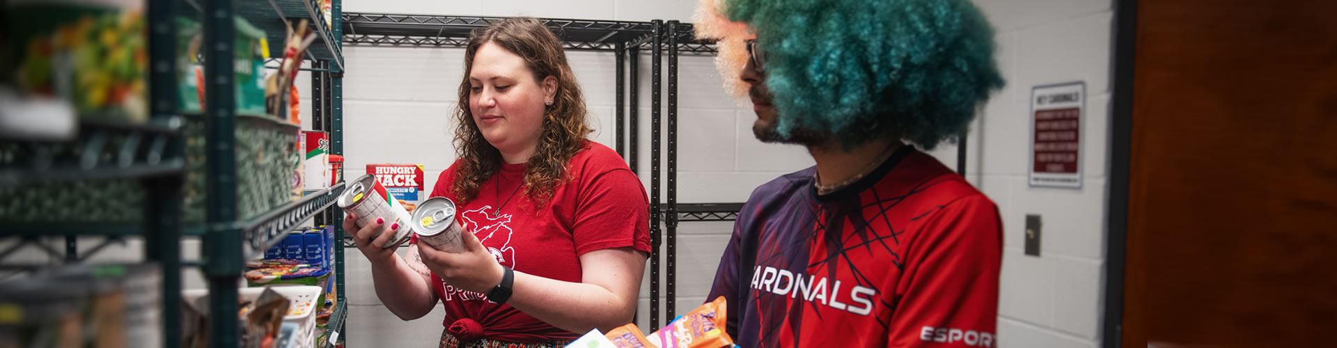 One student holding a box filled with non-perishable food in the Cardinal Food Pantry while the other student add more items to the box.