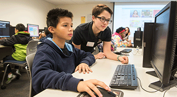 Young male student working at a computer and getting help from another student.